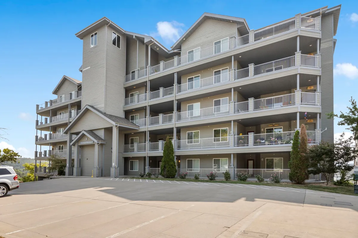 An exterior shot of a modern, multi-story building with balconies, landscaped greenery, and a parking area. The image conveys opportunities for enjoying the outdoors from a private balcony as well as exploring the surrounding landscaped areas.
