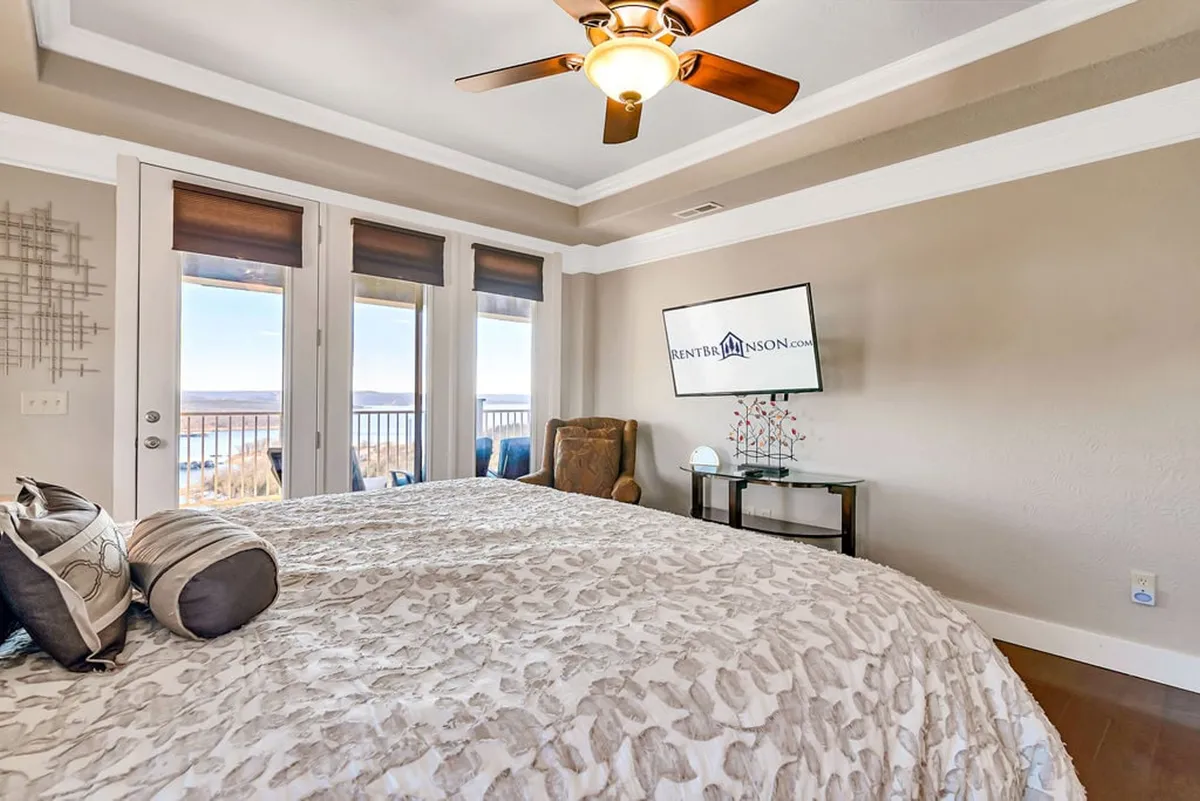 A restful king bedroom with a large bed, plush decorative pillows, a TV, a comfortable chair, and windows offering beautiful views. The space is designed for both relaxation and entertainment.
