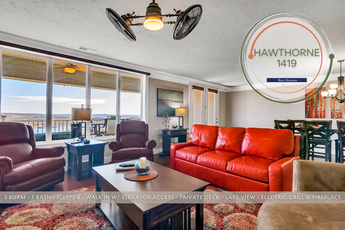 A cozy open-plan living area with a vibrant red sofa and reclining chairs arranged for conversation and comfort, all oriented toward large floor-to-ceiling windows that offer stunning panoramic lake views. The space flows seamlessly into a dining area, and the natural light fills every corner, creating a welcoming ambiance.
