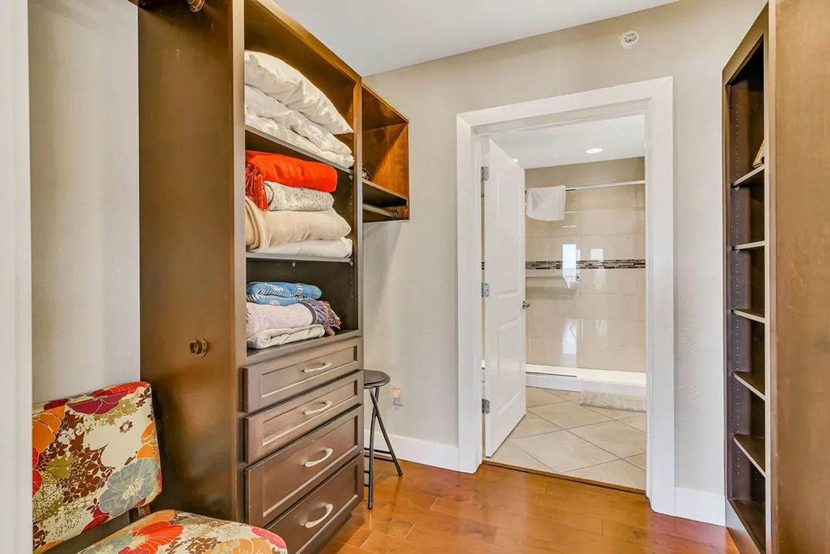 A calm, organized space featuring neatly stacked towels, a colorful chair, and an open door leading to a bathroom. The area is bright and accessible, highlighting thoughtful storage and ease of movement.