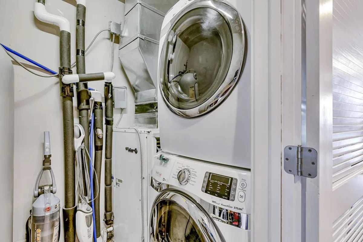 The image shows a tidy, dedicated laundry area equipped with a washer and dryer. Exposed pipes and vents suggest easy access for use, while the overall look is practical and efficient.