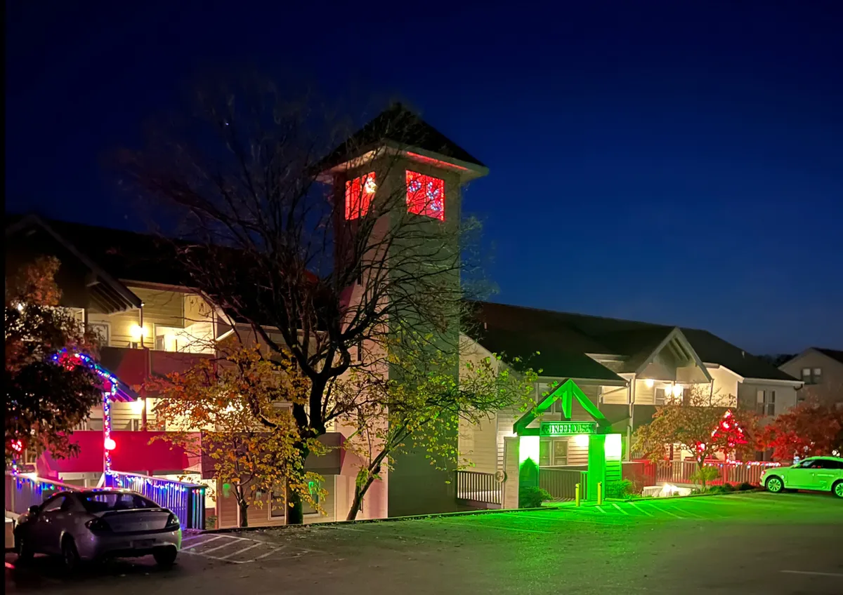 A lively exterior night scene featuring an illuminated clock tower adorned with festive green and red holiday lights and decorations. The surrounding area glows with cheer, evoking the energy of community gatherings and celebrations.