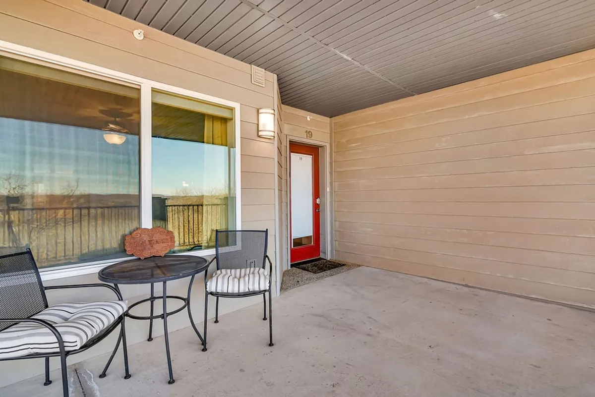 This image features an exterior view of the condo, highlighting a red front door, a circular table with chairs, and a large window that likely faces the lake. The atmosphere is welcoming and hints at moments of enjoying morning coffee or relaxed conversations with a backdrop of sunrise views.